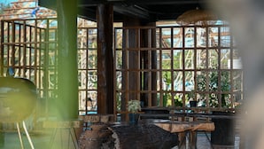 Un espacio de estar al aire libre de estilo rústico con mesas y sillas de madera, un gran tronco como elemento central, y una vista a través de una estructura de celosías de madera.