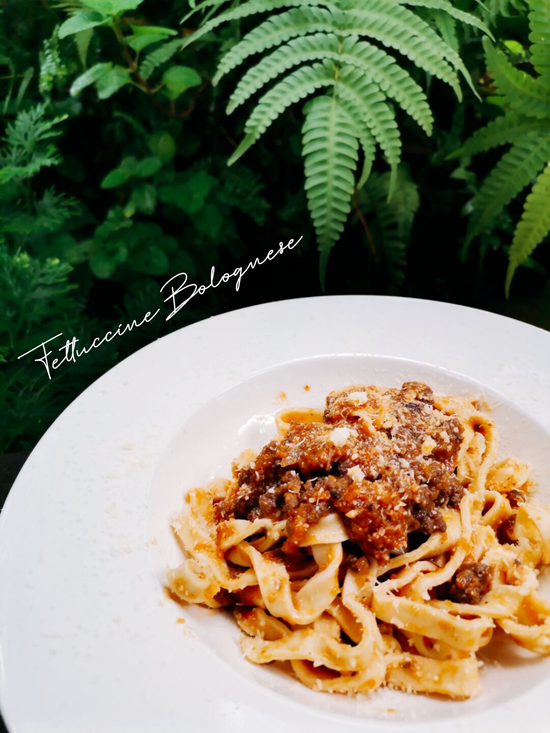 A plate of pasta with a meat-based sauce and grated cheese, set against a backdrop of green foliage.