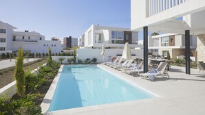 Un área de piscina moderna con sillones de descanso y sombrillas, rodeada de edificios con balcones y un cielo despejado.
