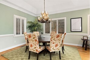Een eetkamer met een ronde tafel, bloemenstoelen, een kroonluchter en een ingelijste kaart aan de muur.