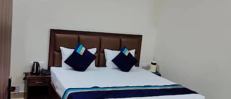 Una cama bien hecha con cabecera de madera, una mesita de noche con un teléfono y una tetera, y una pequeña alfombra en el suelo.