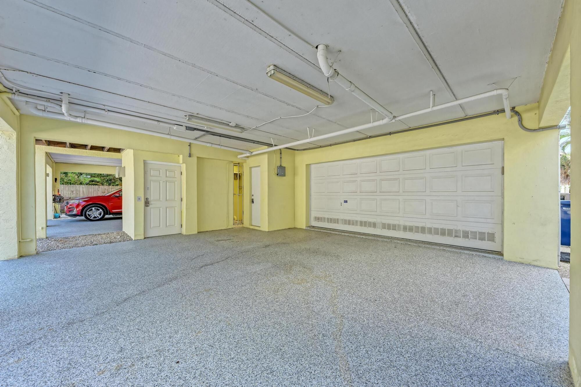 A garage with a red car parked inside, a white garage door, and fluorescent lighting on the ceiling.