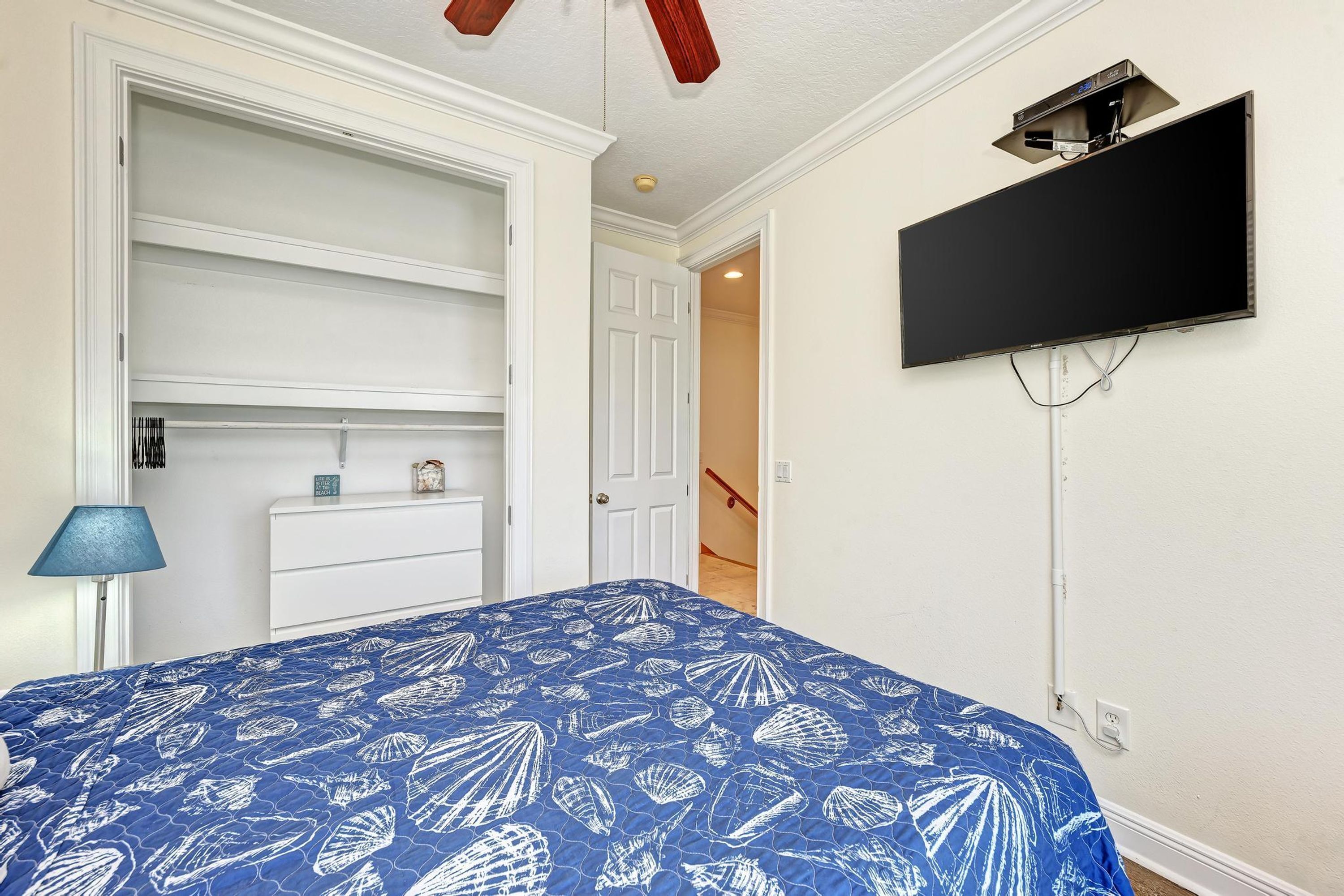 Un dormitorio con una cama, un televisor montado en la pared, una puerta que da a otra habitación y una lámpara en una mesita de noche.