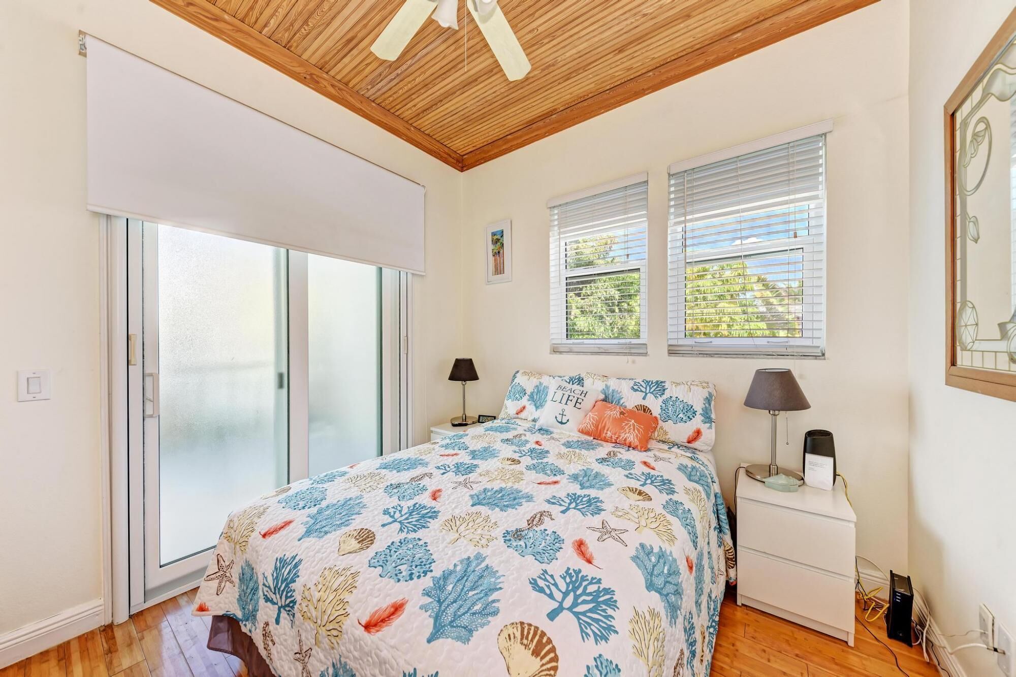A bedroom with a bed, two bedside lamps, a nightstand, a sliding glass door, and two windows with blinds.
