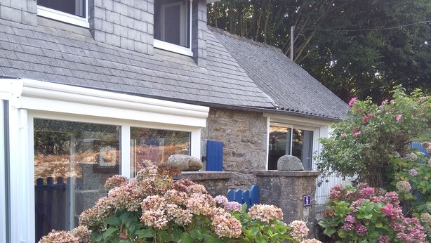 Une maison avec une façade en pierre, un toit gris et un jardin agrémenté de buissons d’hortensias.