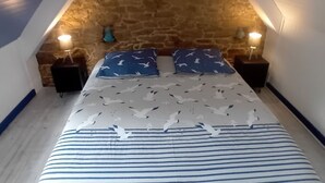Une chambre avec un plafond en pente, un mur en pierre, un lit avec une literie à rayures, deux tables de chevet avec des lampes et un coussin bleu orné d’un motif d’oiseau.
