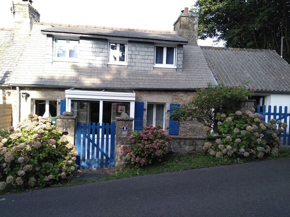 Une maison à deux étages avec une porte bleue, des murs en pierre et des arbustes d’hortensias.