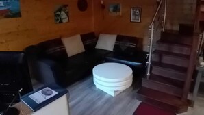 Un salon chaleureux avec un plafond en bois, un canapé noir, un pouf blanc et un escalier menant à un niveau supérieur.