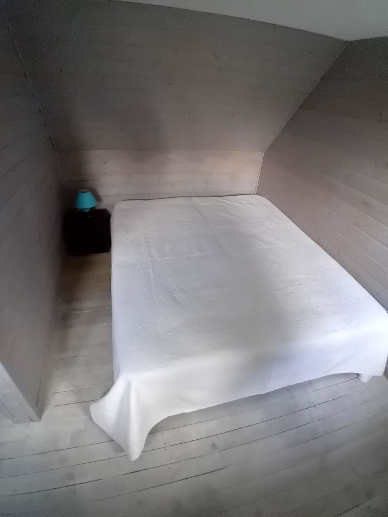 Et lille, svagt oplyst rum med en enkelt seng, et lille bord med en lampe og et mørkt skab.