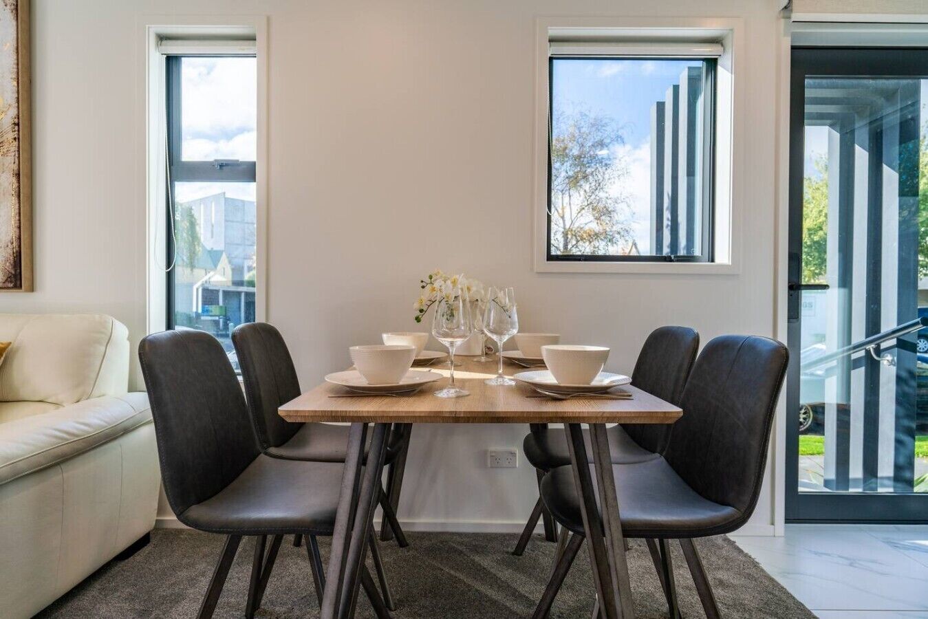 Comedor con una mesa dispuesta para cuatro, dos ventanas y una escalera visible a través del ventanal.