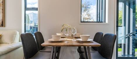 Comedor con una mesa dispuesta para cuatro, dos ventanas y una escalera visible a través del ventanal.