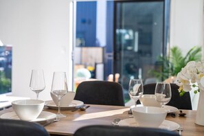 A dining table set with white dishes and glasses, a vase with white flowers, and a view of a cityscape through a large window.
