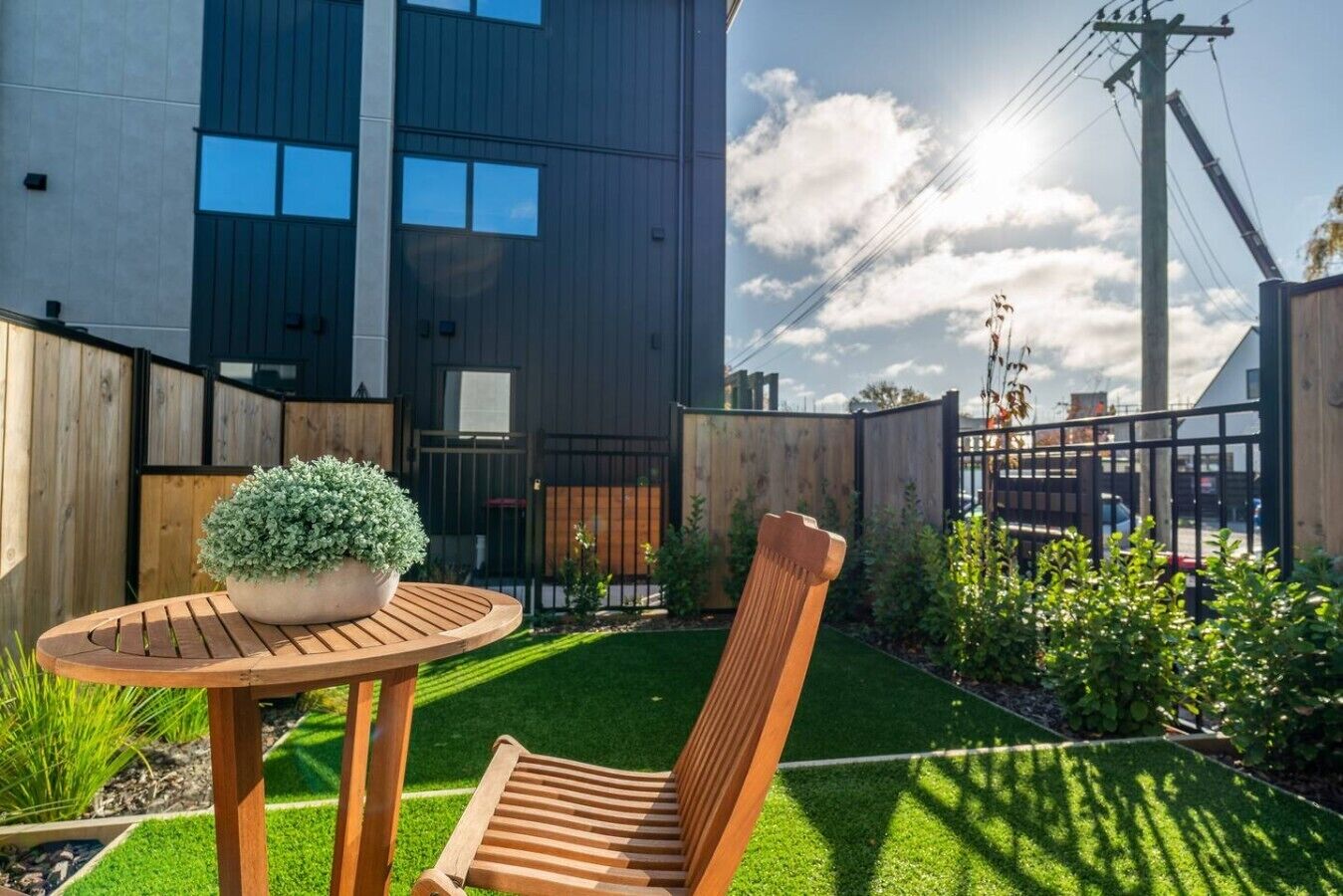 A modern building with a flat roof, a fenced backyard, a wooden table with a potted plant, and a wooden chair.