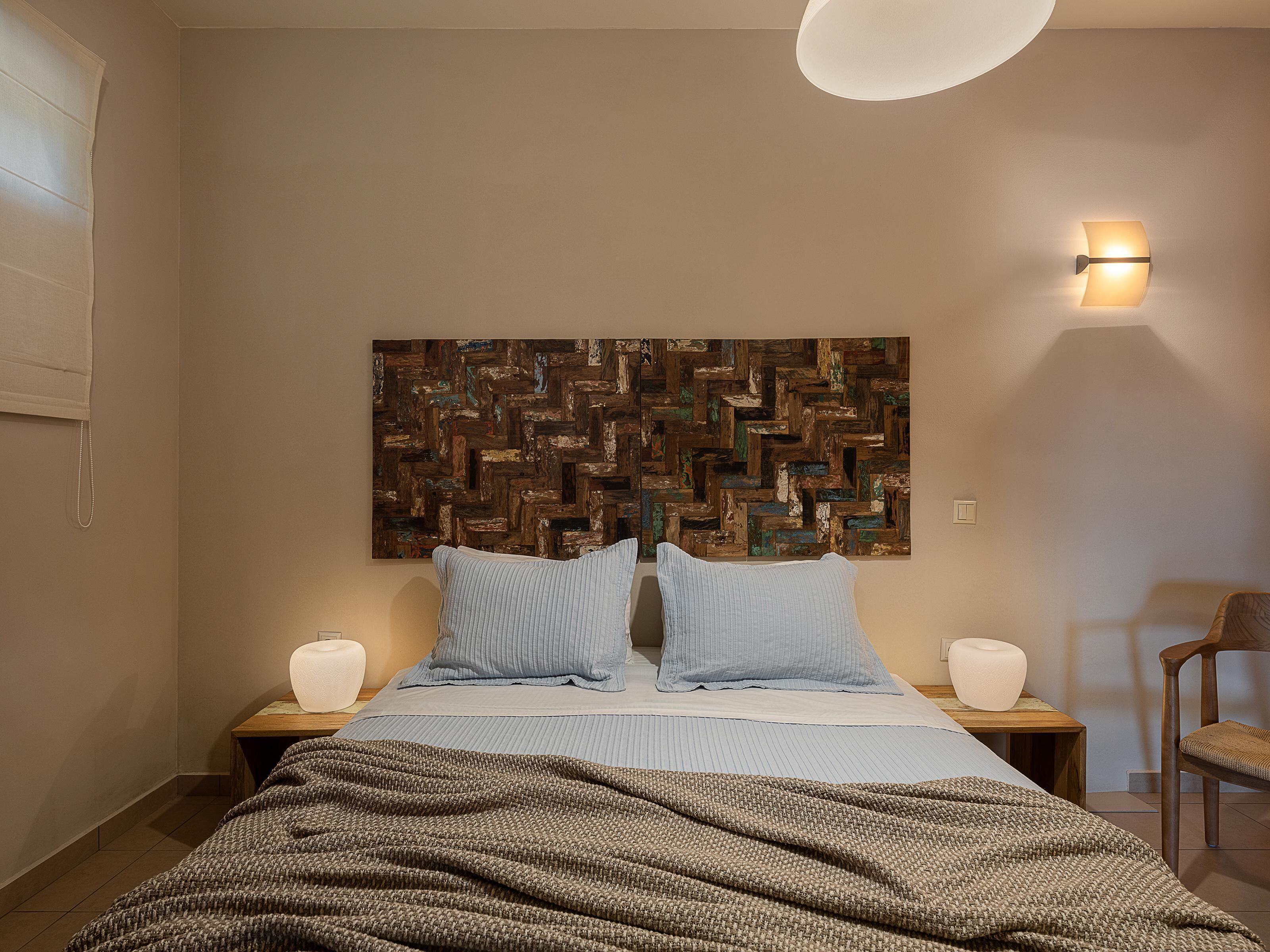 A bedroom with a bed, two bedside tables, a wall-mounted light, a chair, and a textured wall art piece.