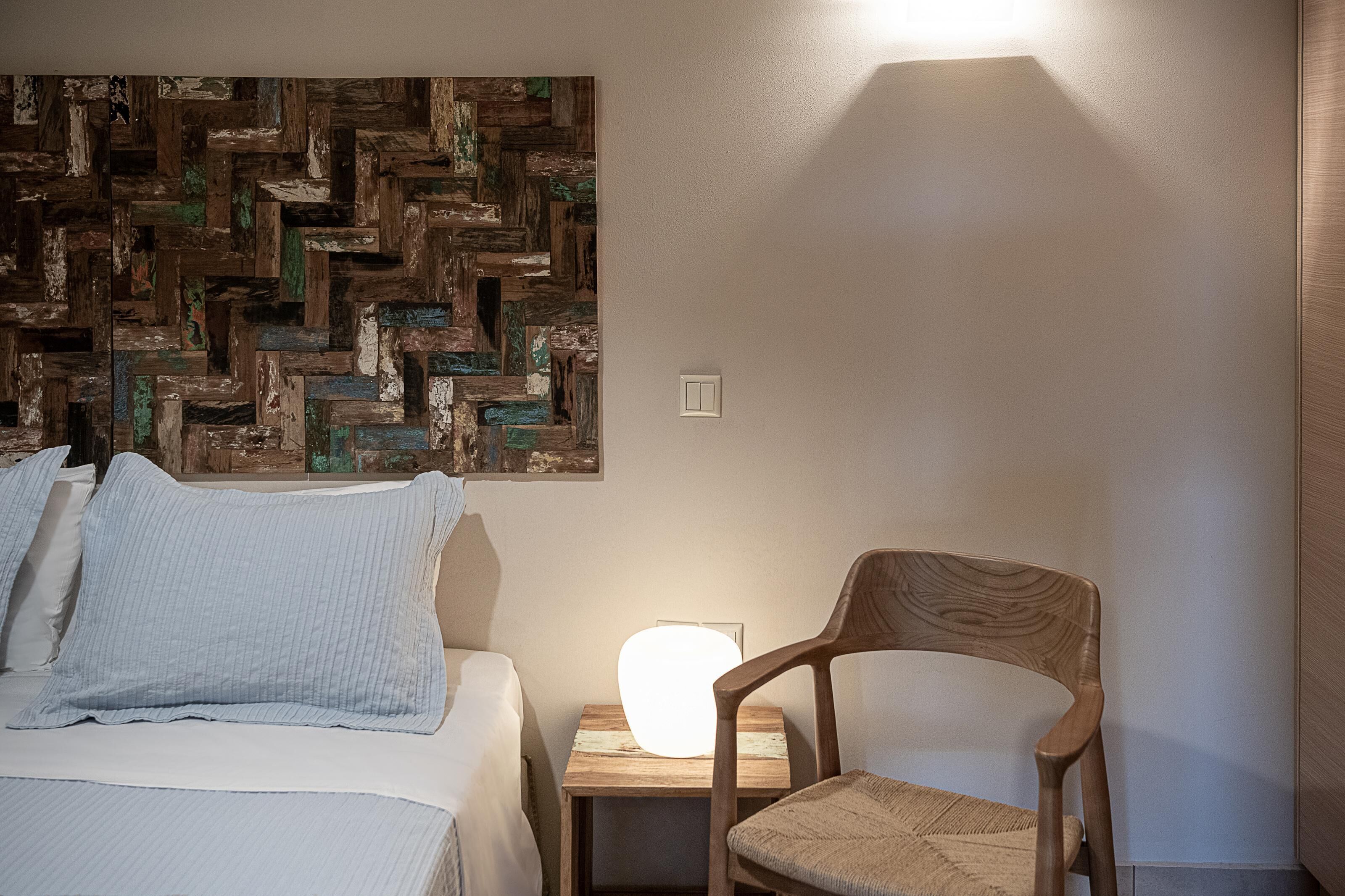 A bedroom with a bed, pillows, a wooden chair, a bedside table with a lamp, and a wall art piece.