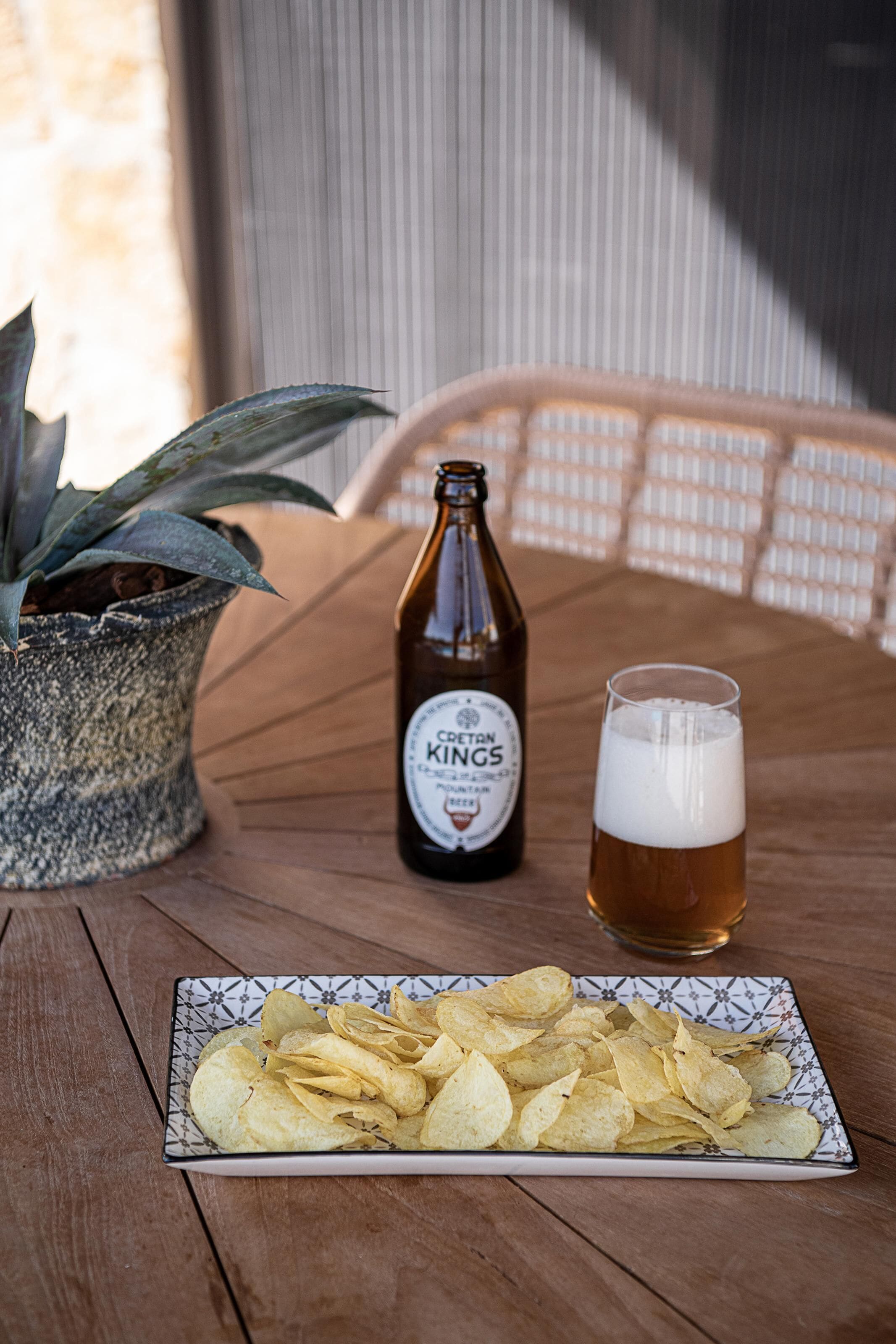 A wooden table with a bottle of beer, a glass of beer, and a plate of chips.