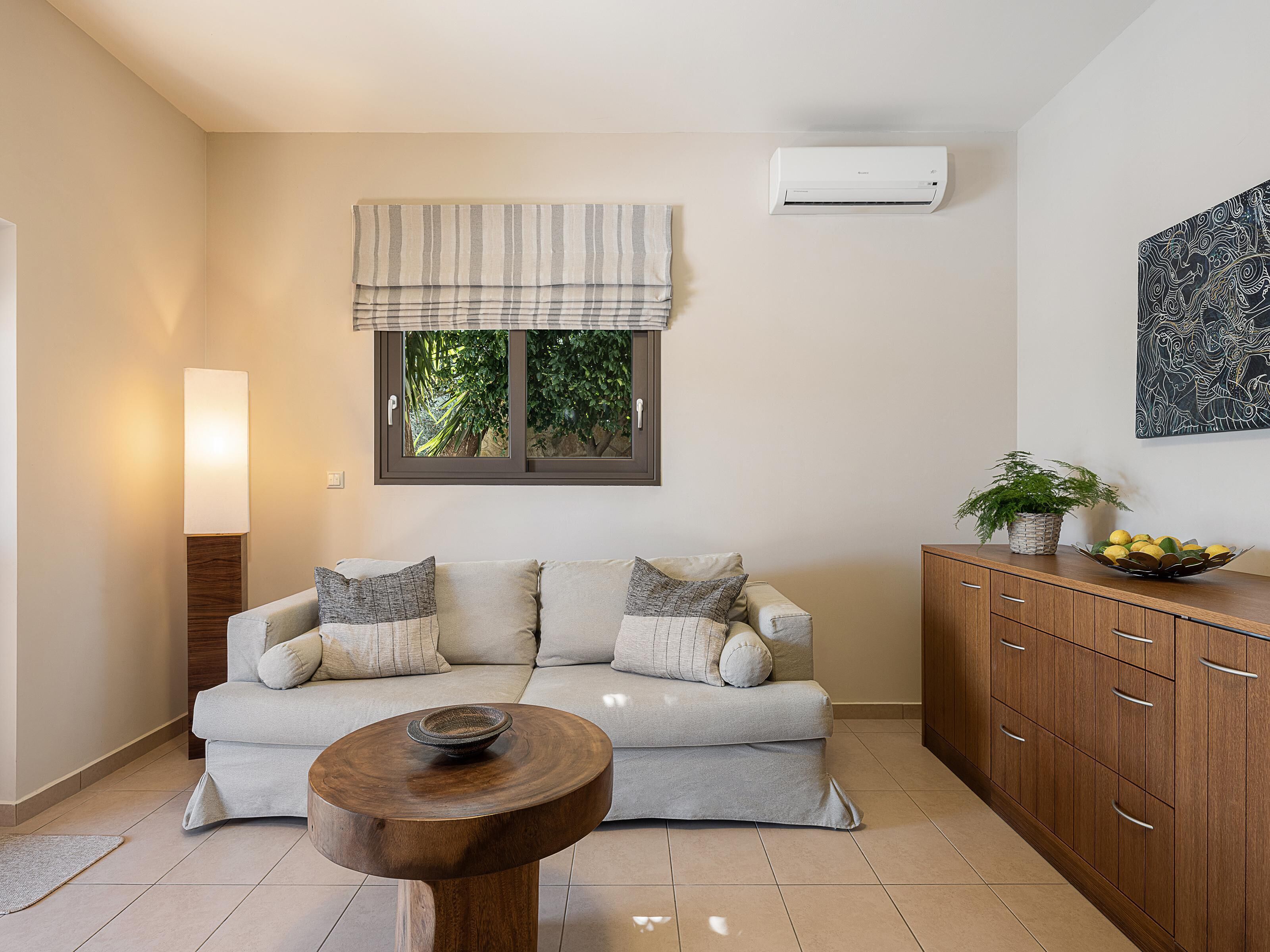 Una sala moderna con un sofá, una mesa de centro de madera, un mueble, un ventanal con vistas a la vegetación y un equipo de aire acondicionado.