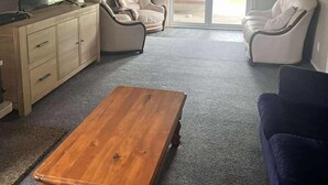 Un salon avec une table basse en bois, deux fauteuils blancs, un canapé beige, un téléviseur posé sur un meuble en bois et une porte-fenêtre coulissante donnant sur l’extérieur.