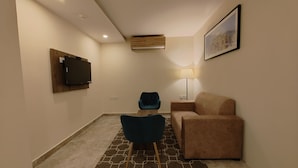 A modern living room with a flat-screen TV mounted on the wall, a blue armchair, a beige sofa, a patterned rug, and a floor lamp. - Regenta Inn Greater Noida (Greater Noida)
