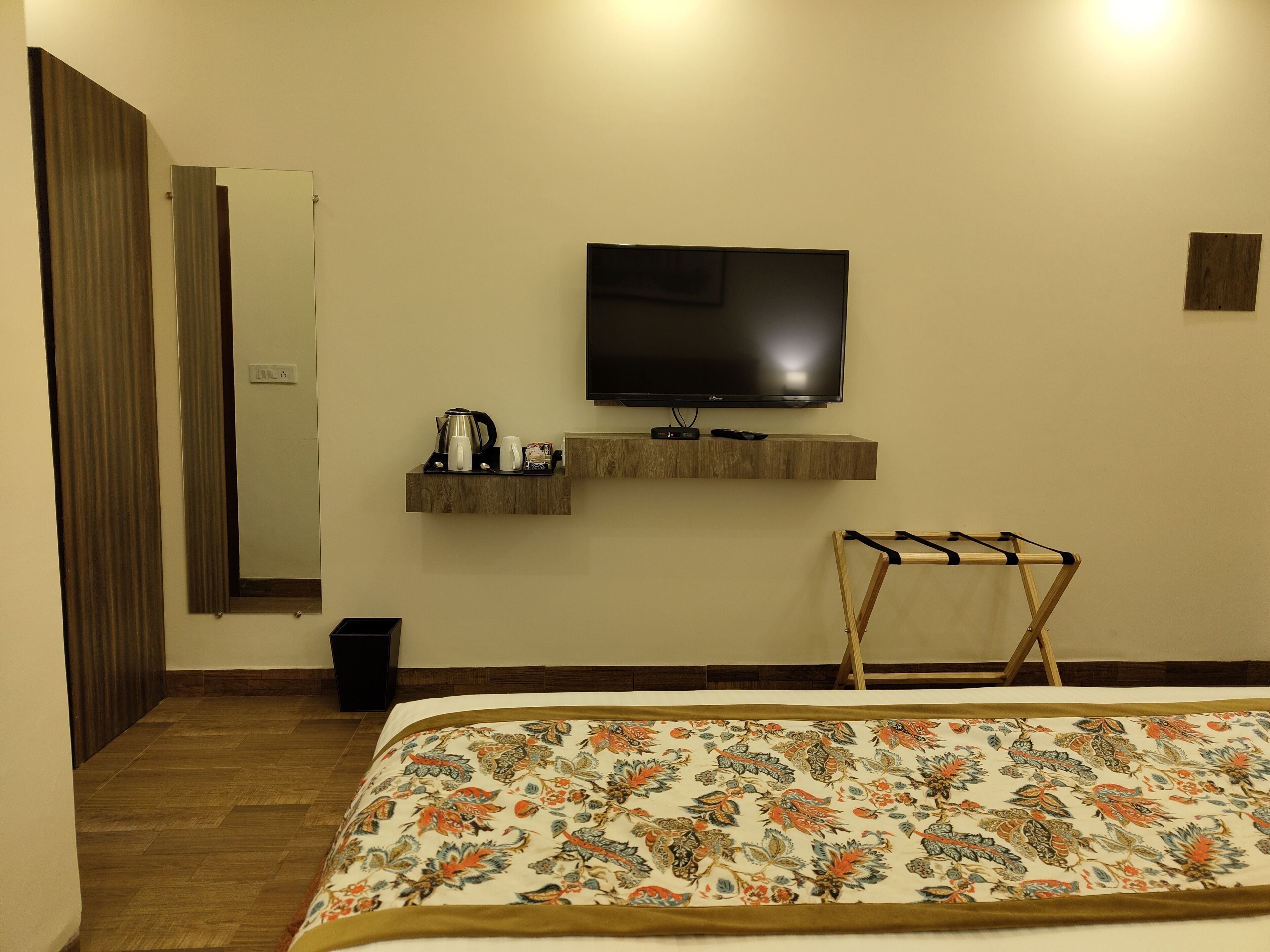 A hotel room with a bed, a TV mounted on the wall, a shelf with a kettle and other items, and a wooden drying rack.