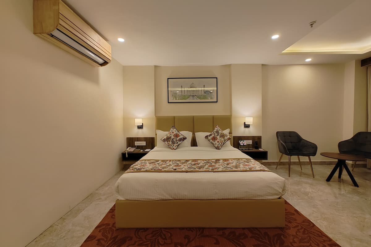 A hotel room with a large bed, two bedside tables, a nightstand, a wall-mounted air conditioner, a framed picture, and a seating area with two chairs and a small table.