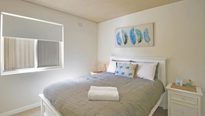 Un dormitorio con cama, mesita de noche, ventana con persianas y una obra de arte en la pared.