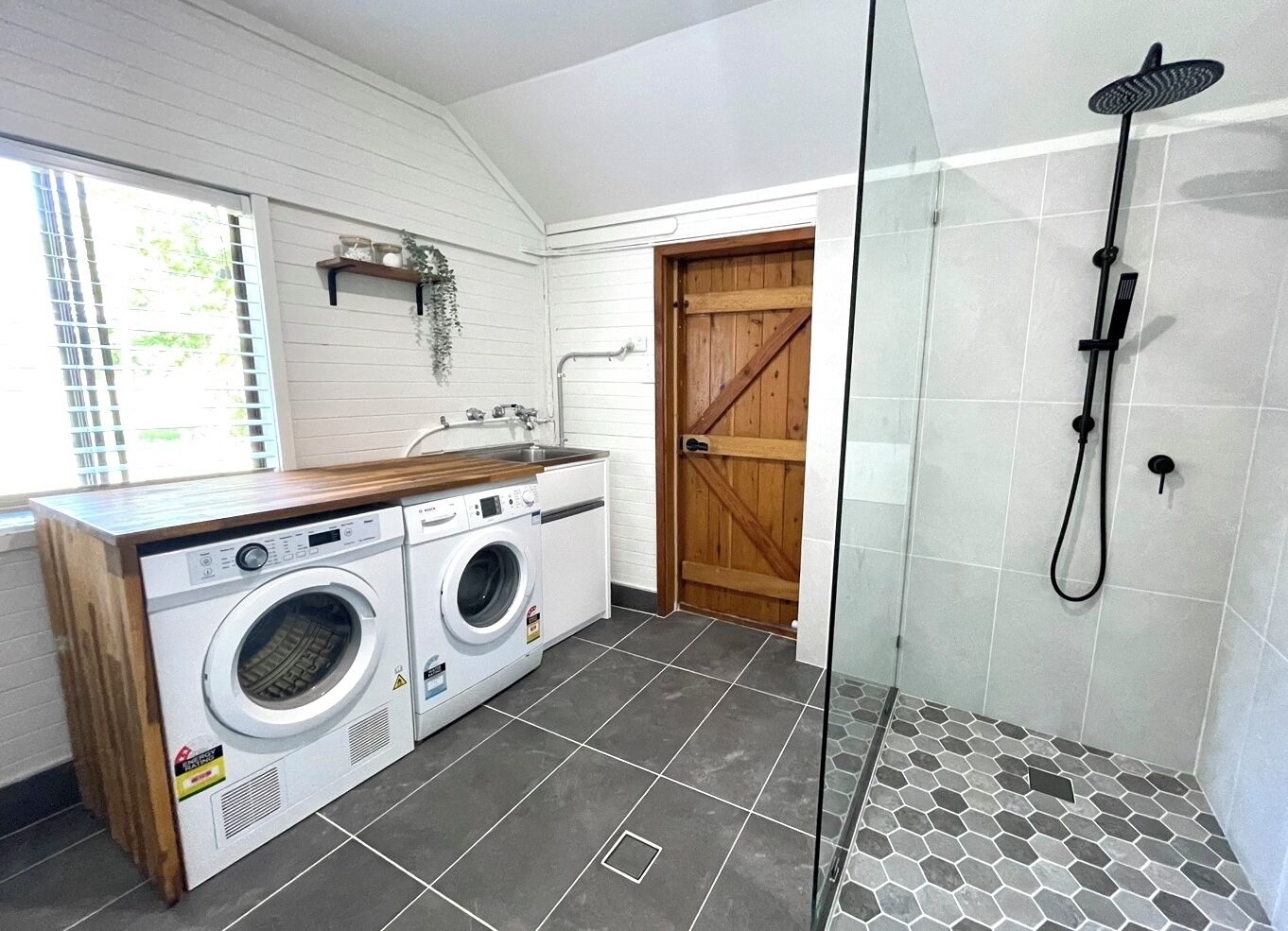 A laundry room with a washing machine and dryer, a wooden countertop, a glass shower enclosure, and a tiled floor.