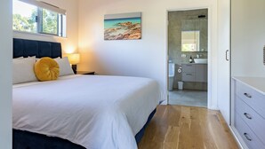 A bedroom with a bed, a nightstand, a lamp, a painting, a bathroom with a sink and mirror, and wooden flooring.
