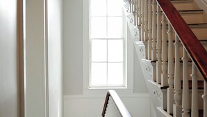 Una escalera con balaustres blancos y barandilla de madera que lleva a un rellano con una ventana.
