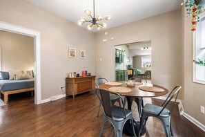 A dining area with a round table, chairs, and a chandelier. There is a sofa and a cabinet in the adjacent room.