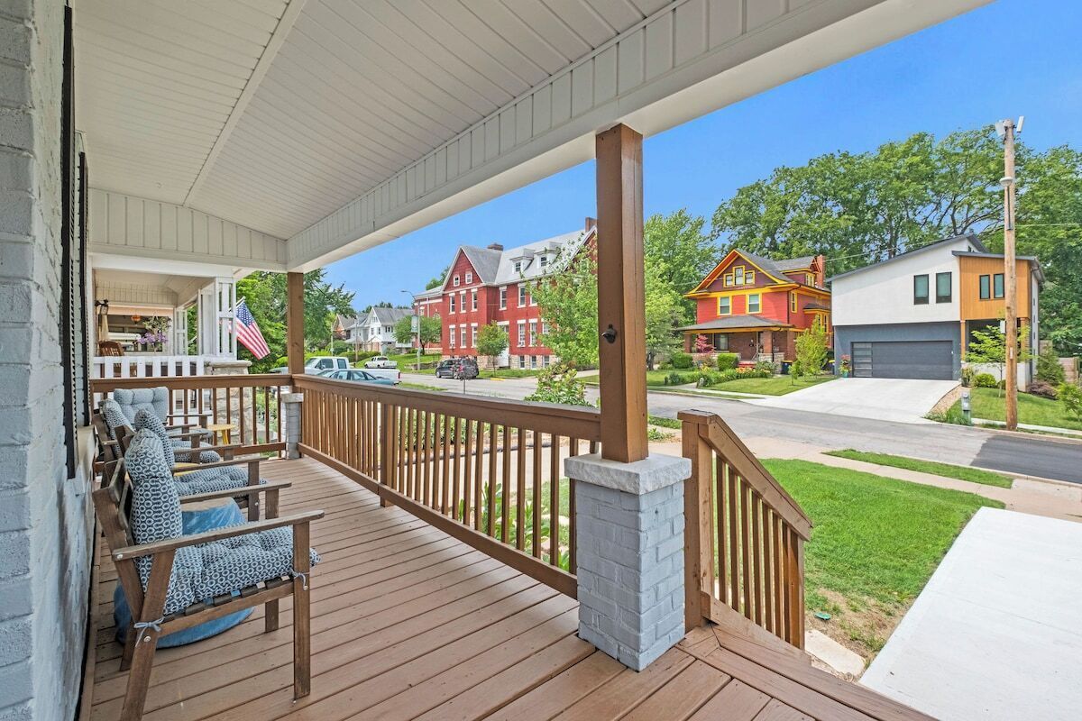 A covered porch with wooden railings and seating, overlooking a street with houses and a clear sky.
