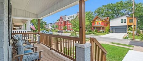 A covered porch with wooden railings and seating, overlooking a street with houses and a clear sky.