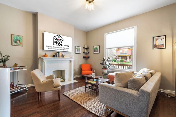 A living room with a fireplace, a sofa, a chair, and a coffee table. There is a sign on the wall and a window with a view of buildings outside.