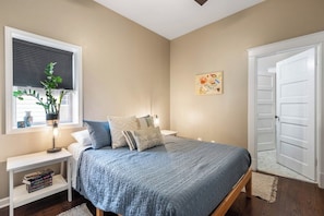 A bedroom with a bed, bedside tables, a window with blinds, a door, and a wall-mounted artwork.