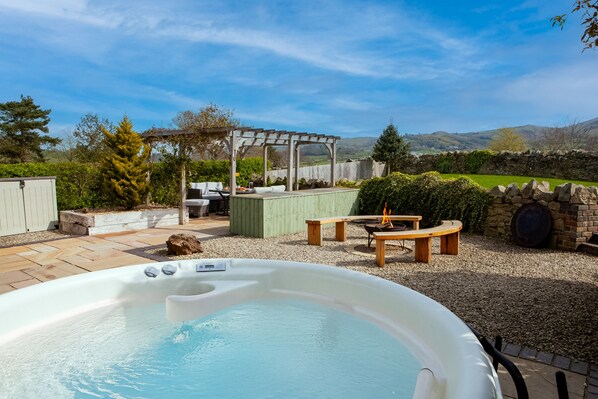 A hot tub with a wooden table and chairs, surrounded by a stone wall and a pergola.