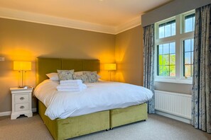 A neatly made bed with white linens and a green headboard, a bedside table with a lamp, and a window with curtains.