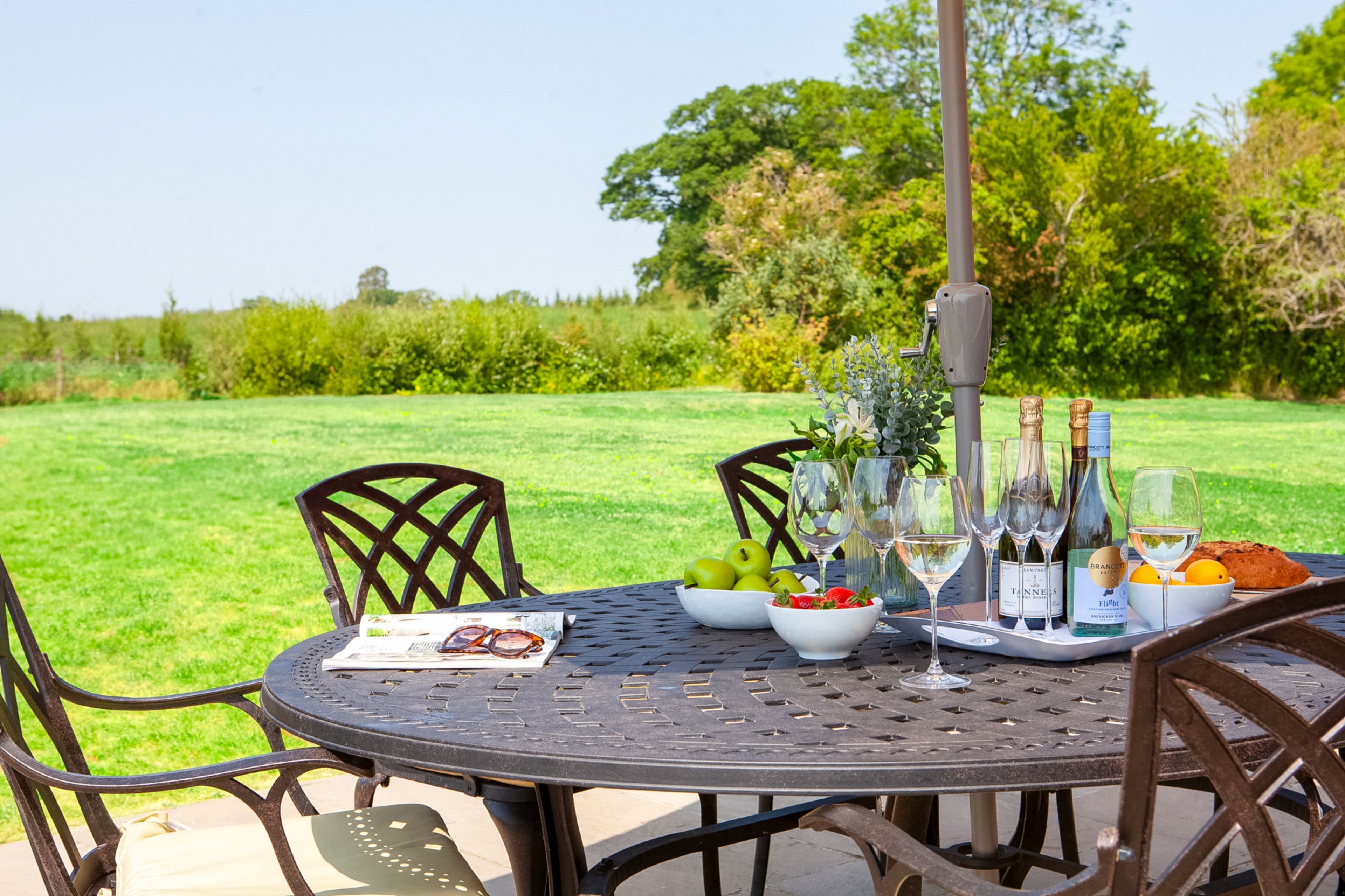 An outdoor dining table with chairs, a bottle of wine, glasses, and a bowl of fruit.