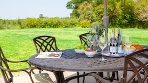 Mesa de jantar ao ar livre com cadeiras, uma garrafa de vinho, copos e uma cesta de frutas.