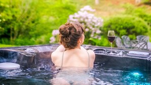 En person i ett bubbelbad med ett glas rött vin, omgiven av frodig grönska.