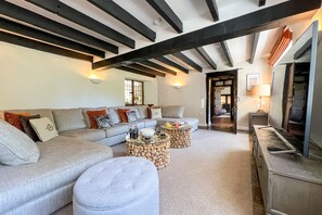 A living room with a sofa, ottoman, and coffee table, wooden beams on the ceiling, and a door leading to another room.