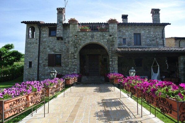 A stone building with an arched entrance, a balcony with potted plants, and a pathway lined with flower beds.