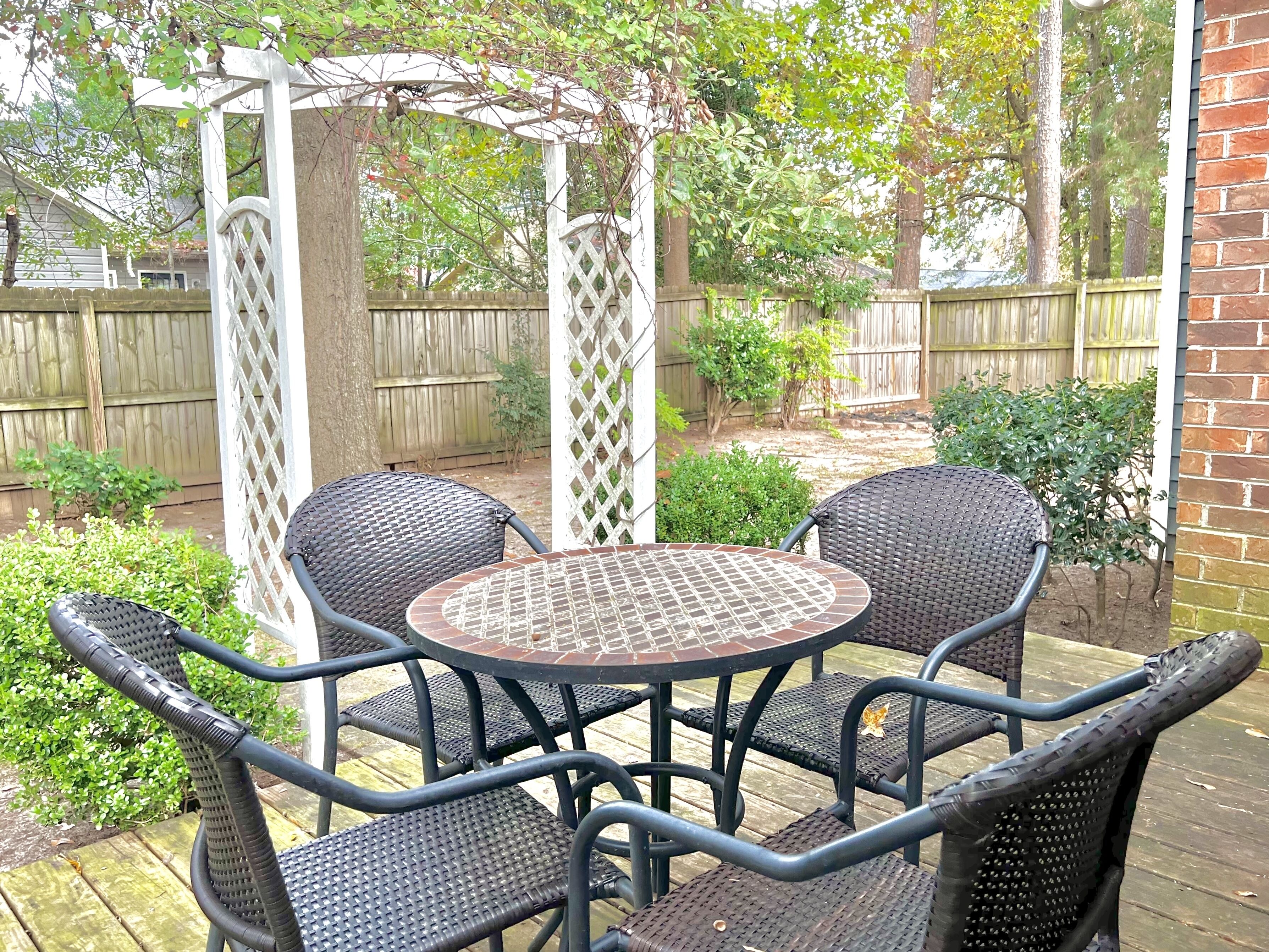 A patio area with a round table and four chairs, surrounded by a wooden deck and a white lattice fence.