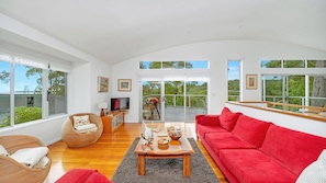 A spacious living room with a red sofa, a wicker chair, a wooden coffee table, and a television. Large windows provide views of the outdoors.