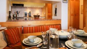 A cozy dining area with a wooden table set for two, a bench with striped cushions, and a kitchen in the background.