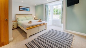 A bedroom with a bed, bedside table, lamp, and a view of the outdoors through a window. - Cherry Orchard Cottage by Interhome (Mevagissey)