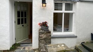 A white building with a stone staircase leading to a door, a window with a white frame, and a potted plant with red flowers. - Cherry Orchard Cottage by Interhome (Mevagissey)
