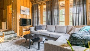 En mysig stuga med träpanel på insidan, en grå soffa, ett svart soffbord, en tv på ett träställ och stora fönster med gardiner.