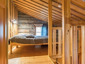 A log cabin bedroom with a lofted bed, wooden beams, and a window with curtains.