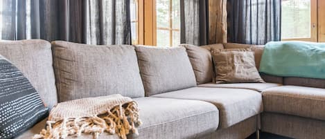 A sectional sofa with a throw blanket and pillows, a small round table, and a view through large windows with curtains.