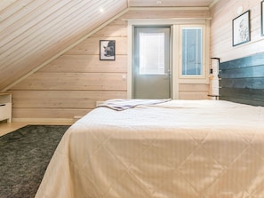 Ein Schlafzimmer mit schrägen Wänden, einem Bett mit gemustertem Überwurf und einer Tür mit Fenster, sowie einem an der Wand befestigten Regal.